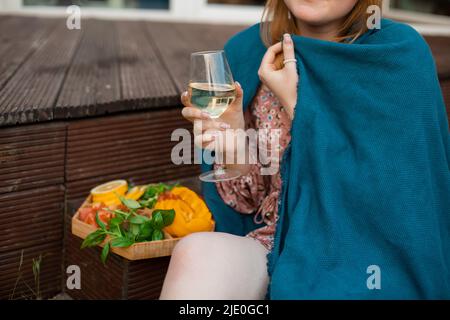 Femme enveloppée de tissu écossais bleu assis sur la terrasse avec un verre de vin blanc et une assiette d'en-cas à proximité. Boire de l'alcool et profiter de l'extérieur Banque D'Images