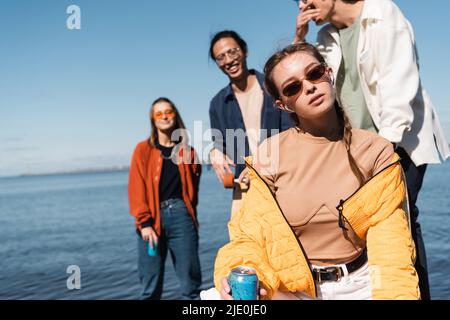 jeune femme en lunettes de soleil tenant le soda can et regardant la caméra près de flou inter-racial amis Banque D'Images