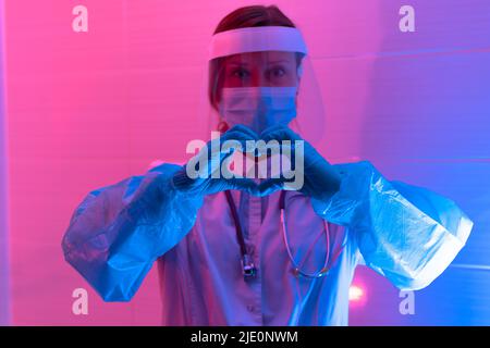 Belle jeune femme médecin avec un stéthoscope dans un masque de protection et des gants dans un laboratoire bio sur fond rouge-bleu montre des signes avec sa main Banque D'Images
