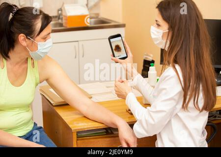 Dermatologue montrant une image rognée de la mole au patient à l'aide d'un appareil moderne utilisant la lumière, l'application de téléphone et l'appareil photo Banque D'Images