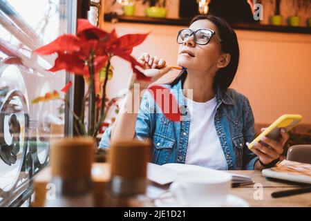 Portrait d'une fille brune portant des lunettes étudiant dans un café tenant un smartphone et un stylo dans ses mains regardant soigneusement par la fenêtre Banque D'Images