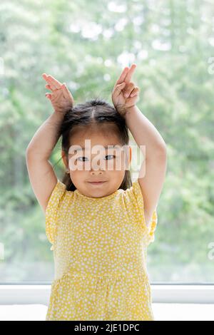 portrait d'une fille asiatique de brunette et de jeune fille en robe jaune montrant des oreilles de lapin près de la fenêtre à la maison Banque D'Images