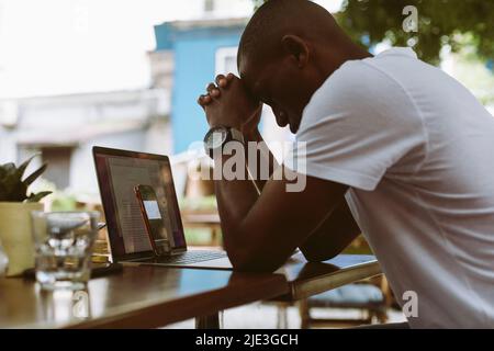 Épuisé, stressé peau sombre homme se penchait la tête sur les mains des articulations près de l'ordinateur portable et du smartphone. Conférence en ligne dans le café Banque D'Images