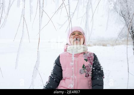 Caucasien mignon jeune fille avec la bouche ouverte attrapant des flocons de neige avec la langue avec les yeux ouverts avec des brindilles d'arbre et champ neigeux en arrière-plan. Hiver Banque D'Images