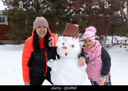 Une jeune fille souriante et une femme d'âge moyen embrassant un bonhomme de neige pendant la journée tout en se promenant dans le parc avec des arbres en arrière-plan. Les parents passent du temps avec Banque D'Images