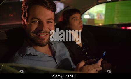 Homme heureux en voiture, assis à l'arrière la nuit après le travail, portrait souriant tenant le smartphone. Deux personnes en taxi Banque D'Images