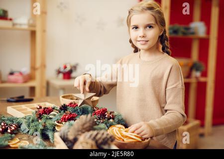 Portrait d'une petite fille souriante avec des tresses debout au bureau avec des matériaux décoratifs et décorant la couronne de Noël avec le jouet Banque D'Images