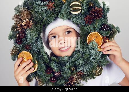 Portrait d'une petite fille souriante regardant à travers la couronne de Noël décoré de cônes, de tranches d'orange et de petites boules Banque D'Images