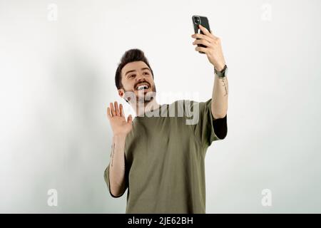 Portrait d'un homme gai portant un t-shirt kaki posé isolé sur fond blanc prenant le selfie ou ayant un appel vidéo et la main ondulée. Enregistrement vlo Banque D'Images