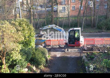 Workman utilise une minipelle pour creuser une chaussée (trottoir) par une route dans une zone résidentielle Banque D'Images