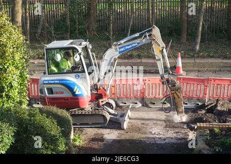 Workman utilise une minipelle pour creuser une chaussée (trottoir) par une route dans une zone résidentielle Banque D'Images