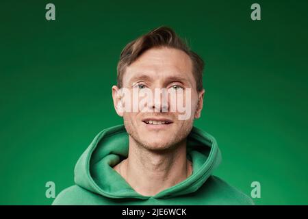 Vue de face gros plan d'un homme caucasien adulte regardant l'appareil photo avec le sourire tout en portant un sweat à capuche sur fond vert vif Banque D'Images