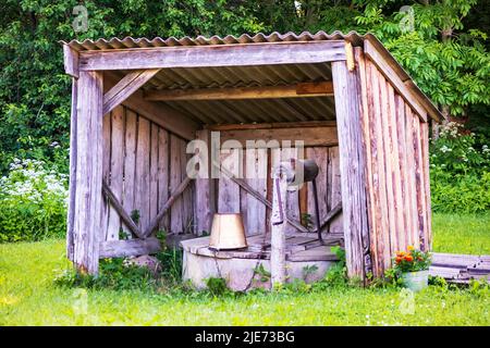 Un ancien puits en bois avec un seau en métal dans une herbe verte avec une maison en bois Banque D'Images