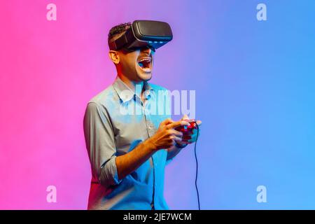 Vue latérale de l'homme excité en chemise dans des lunettes VR jouant à un jeu vidéo, en utilisant un simulateur pour l'éducation, hurler émotionnellement. Studio d'intérieur isolé sur fond de néon coloré. Banque D'Images
