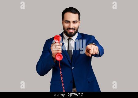 Homme positif pointant le doigt vers l'appareil photo, tenant un téléphone fixe rouge vintage, intéressé par des appareils rétro, répondant aux appels, portant un costume de style officiel. Prise de vue en studio isolée sur fond gris. Banque D'Images