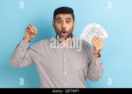 Un homme d'affaires choqué tient montrant des billets en dollars et des pièces d'or de crypto-monnaie, transférant de l'argent dans des bitcoins numériques, en portant une chemise rayée. Studio d'intérieur isolé sur fond bleu. Banque D'Images