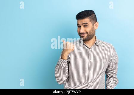 Portrait d'un homme à barbe agréable à regarder pointant le pouce de côté à l'espace de copie, montrant l'espace pour la publicité, portant une chemise rayée. Studio d'intérieur isolé sur fond bleu. Banque D'Images