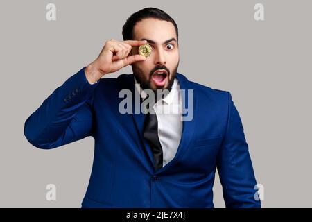 Portrait d'homme d'affaires barbu couvrant l'œil avec un bitcoin doré, regardant l'appareil photo avec une expression choquée, portant un costume de style officiel. Prise de vue en studio isolée sur fond gris. Banque D'Images