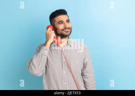 Un homme d'affaires heureux avec une barbe parlant téléphone fixe tenant dans le combiné main, regardant l'appareil photo avec un sourire crasseux, portant une chemise rayée. Studio d'intérieur isolé sur fond bleu. Banque D'Images