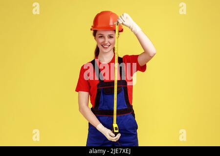 Portrait de femme constructeur tenant la roulette pour mesurer, regardant la caméra avec l'expression du visage satisfait, portant des combinaisons et un casque de protection. Studio d'intérieur isolé sur fond jaune. Banque D'Images