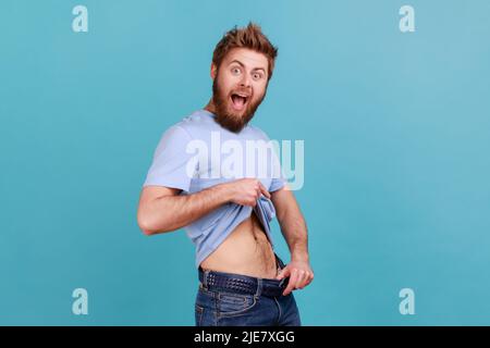 Portrait de l'homme barbu ayant des points de volonté à la taille mince dans le grand pantalon, perte de poids réussie, gardant l'alimentation et aller pour le sport. Studio d'intérieur isolé sur fond bleu. Banque D'Images