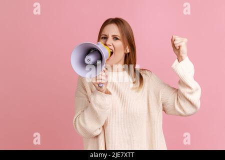 Portrait d'une femme blonde sérieuse et concentrée manifestant, haussé du bras et criant au mégaphone, regardant la caméra, portant un chandail blanc. Studio d'intérieur isolé sur fond rose. Banque D'Images