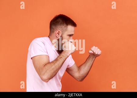 Vue latérale de l'homme barbu combattant agressif tenant les poings serrés prêts à la boxe, entraîneur d'art martial, autodéfense, portant un T-shirt rose. Studio d'intérieur isolé sur fond orange. Banque D'Images