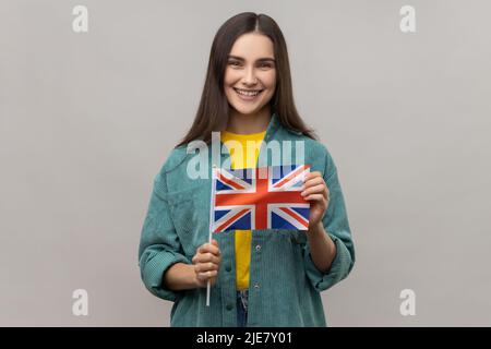 Portrait d'une femme ravie tenant le drapeau britannique, regardant l'appareil photo avec un sourire crasseux, patriote, portant une veste de style décontracté. Prise de vue en studio isolée sur fond gris. Banque D'Images