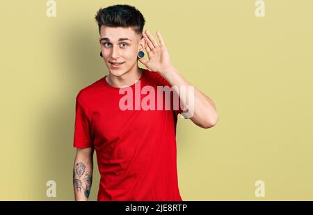 Jeune garçon caucasien avec des oreilles dilatation portant un t-shirt rouge décontracté souriant avec la main sur l'oreille écouter et entendre à la rumeur ou des potins. La surdité concresse Banque D'Images