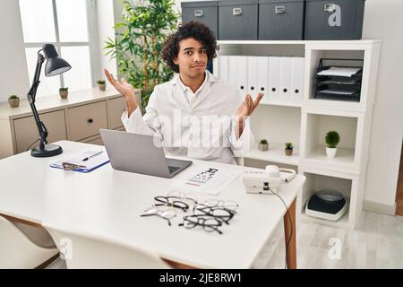 Homme hispanique avec cheveux bouclés travaillant au bureau d'opticien criant et criant à voix haute à côté de la main sur la bouche. Concept de communication. Banque D'Images