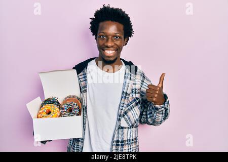 Jeune homme afro-américain tenant une boîte de beignets colorés et savoureux souriant heureux et positif, pouce vers le haut faisant excellent et signe d'approbation Banque D'Images