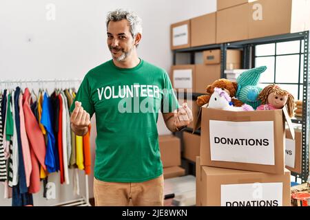 Homme hispanique d'âge moyen portant le t-shirt de volontaire à des dons stand faire le geste d'argent avec les mains, demandant le paiement de salaire, millionnaire affaires Banque D'Images