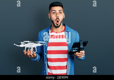 Jeune homme hispanique avec barbe utilisant un drone avec télécommande peur et choqué avec l'expression surprise et stupéfaite, peur et visage excité. Banque D'Images