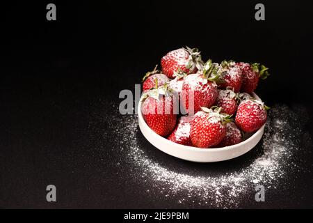 Fraises dans une assiette de sucre en poudre sur fond noir, gros plan Banque D'Images