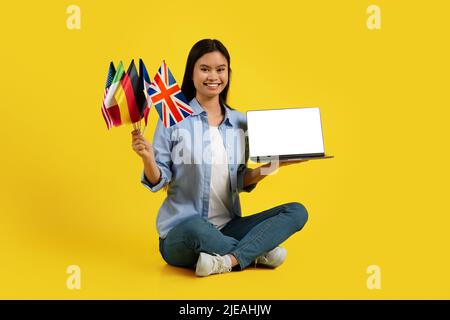 Joyeuse femme asiatique millénaire enseignant, tuteur avec beaucoup de drapeaux différents est assis sur le sol, montre ordinateur portable Banque D'Images