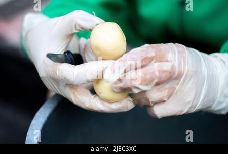 Les mains de la femme en gants blancs pelent de nouvelles pommes de terre brutes, en gros plan. Nettoyage des pommes de terre biologiques. Détail des mains de femmes épluchant la pomme de terre jaune fraîche avec le kitc Banque D'Images