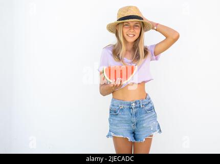 Jeune jolie fille portant un chapeau de paille tenant une tranche de pastèque dans les mains et souriant heureux, sur fond blanc dehors. Concept d'été. Banque D'Images