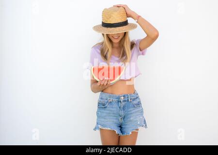 Jeune jolie fille portant un chapeau de paille tenant une tranche de pastèque dans les mains et souriant heureux, sur fond blanc dehors. Concept d'été. Banque D'Images