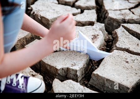 La fille abaisse le bateau en papier sur le sol sec et fissuré. Crise de l'eau et changement climatique concept. Réchauffement de la planète Banque D'Images