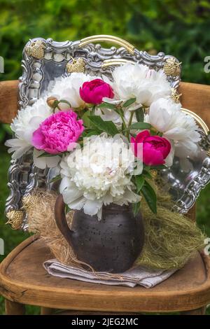 Bouquet de pivoines blanches et roses dans une carafe sur une chaise et sur fond de plateaux argentés. Banque D'Images