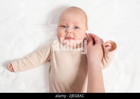 La main de la mère nettoie les oreilles de bébé fille couché sur une feuille blanche avec un coton-tige Banque D'Images