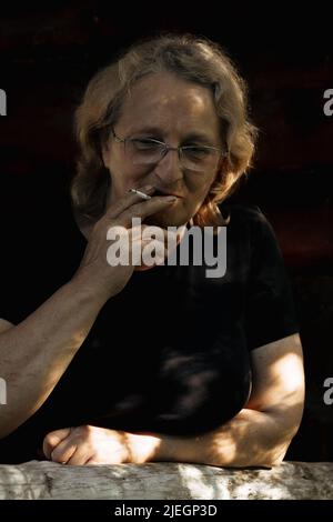 Portrait d'une femme âgée en lunettes avec une cigarette en gros plan sur un fond naturel. Lumière du coucher du soleil sur le visage de l'aîné. Photo de haute qualité Banque D'Images