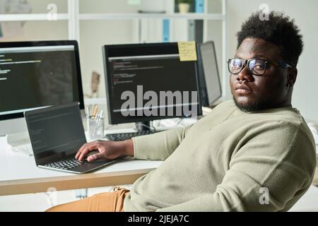 Un programmeur africain sérieux regarde la caméra tout en étant assis près d'un écran d'ordinateur avec un script à l'écran Banque D'Images