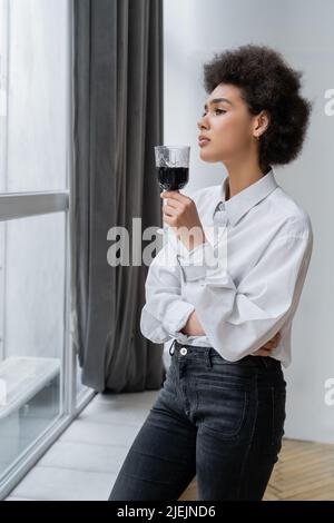 triste femme afro-américaine tenant un verre de vin rouge et regardant la fenêtre Banque D'Images