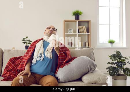 Homme malade qui a la grippe ou le froid assis sur le canapé à la maison, tenant le papier de soie et les éternuements Banque D'Images