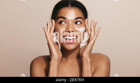 Une femme souriante et heureuse applique une crème hydratante pour le visage, nourrit sa peau, se tient sur fond marron Banque D'Images