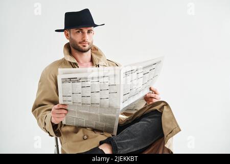 Jeune homme beau dans un manteau de tranchée et chapeau lisant le journal assis sur une chaise sur fond blanc. Concept rétro et détective Banque D'Images