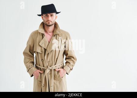 Jeune homme dans un manteau de tranchée et chapeau sur fond blanc avec espace de copie. Concept rétro et détective Banque D'Images