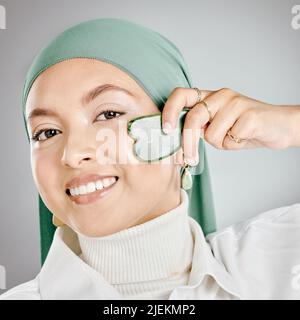 Portrait d'une heureuse belle femme musulmane utilisant un gua sha sur son visage contre un fond gris de studio. Jeune femme portant un hijab et la faisant Banque D'Images