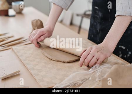 Mains de jeunes artisans avec broche de roulement en bois aplatissant l'argile à bord tout en travaillant sur de nouveaux produits d'argile à vendre en atelier Banque D'Images